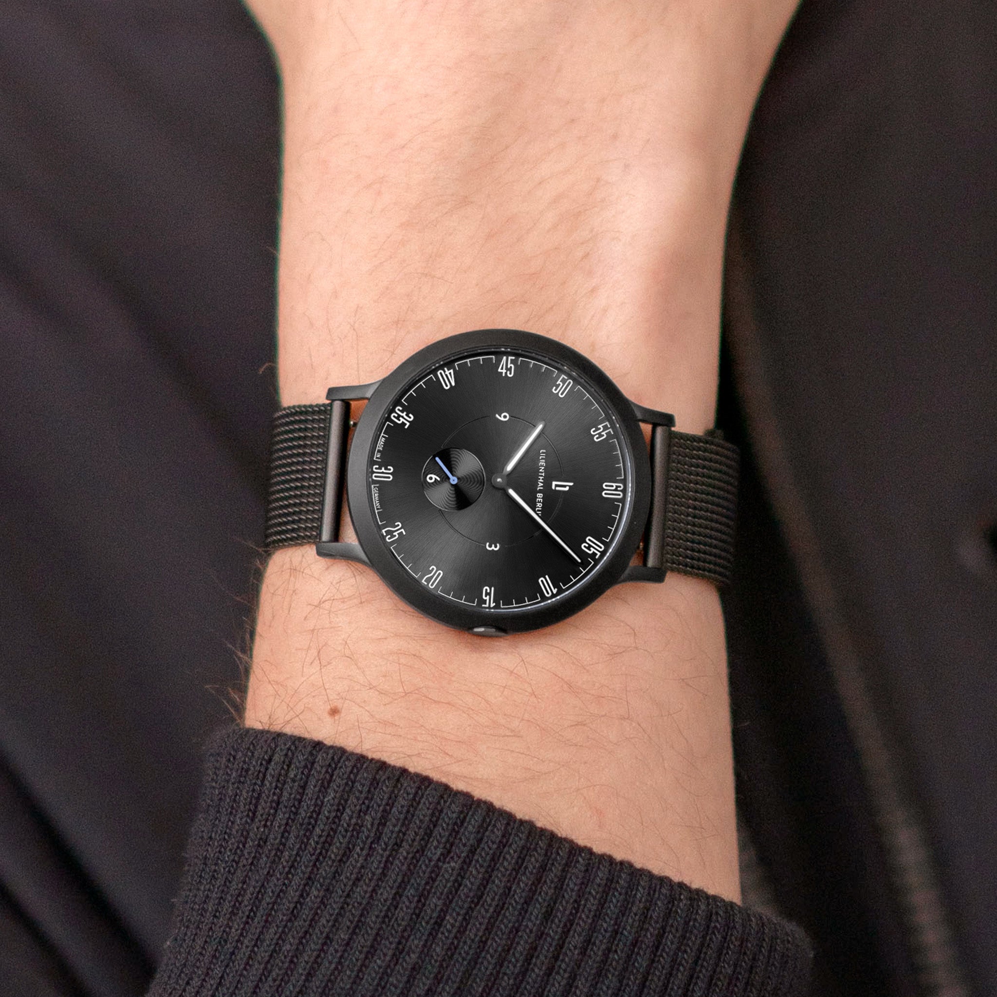 Minimalist black wristwatch with mesh strap on person's wrist, modern analog dial design.