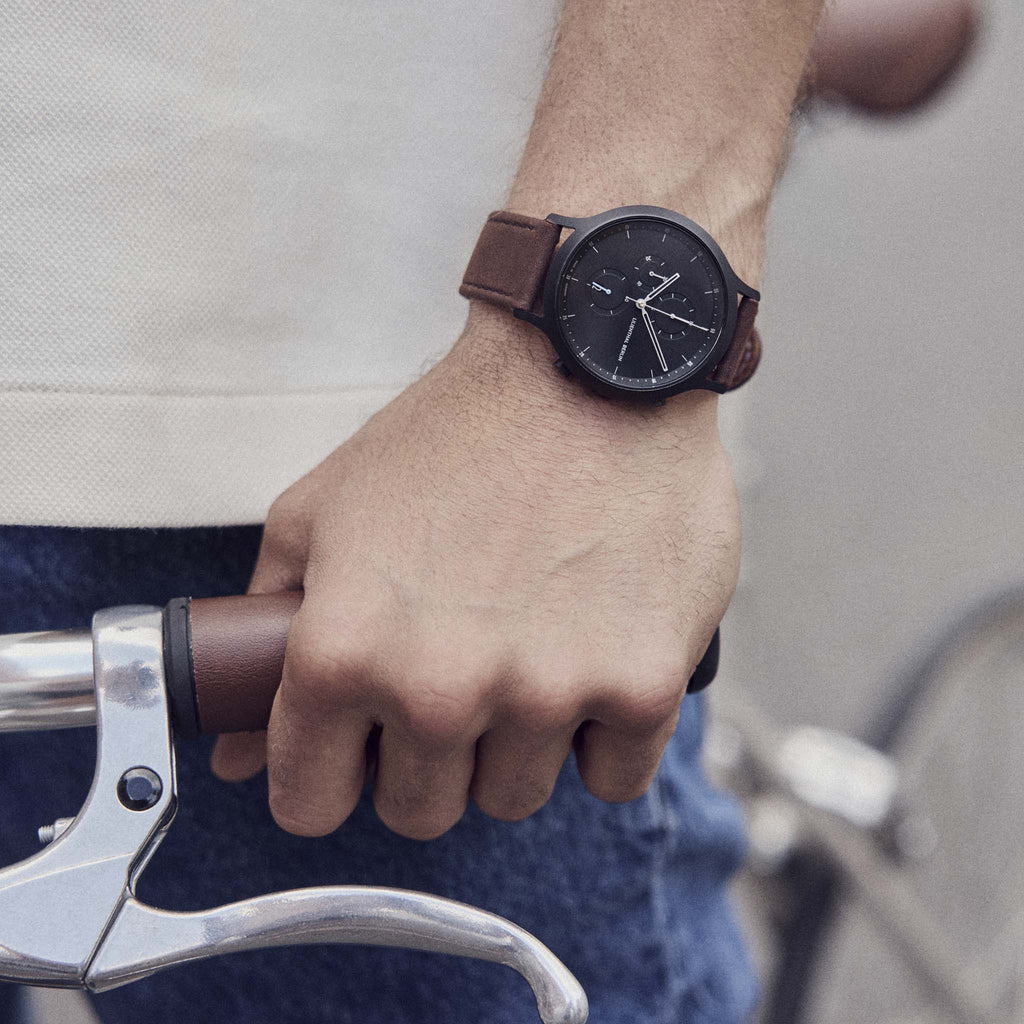 Man wearing black dial wristwatch with brown leather strap, hand on bicycle handlebar