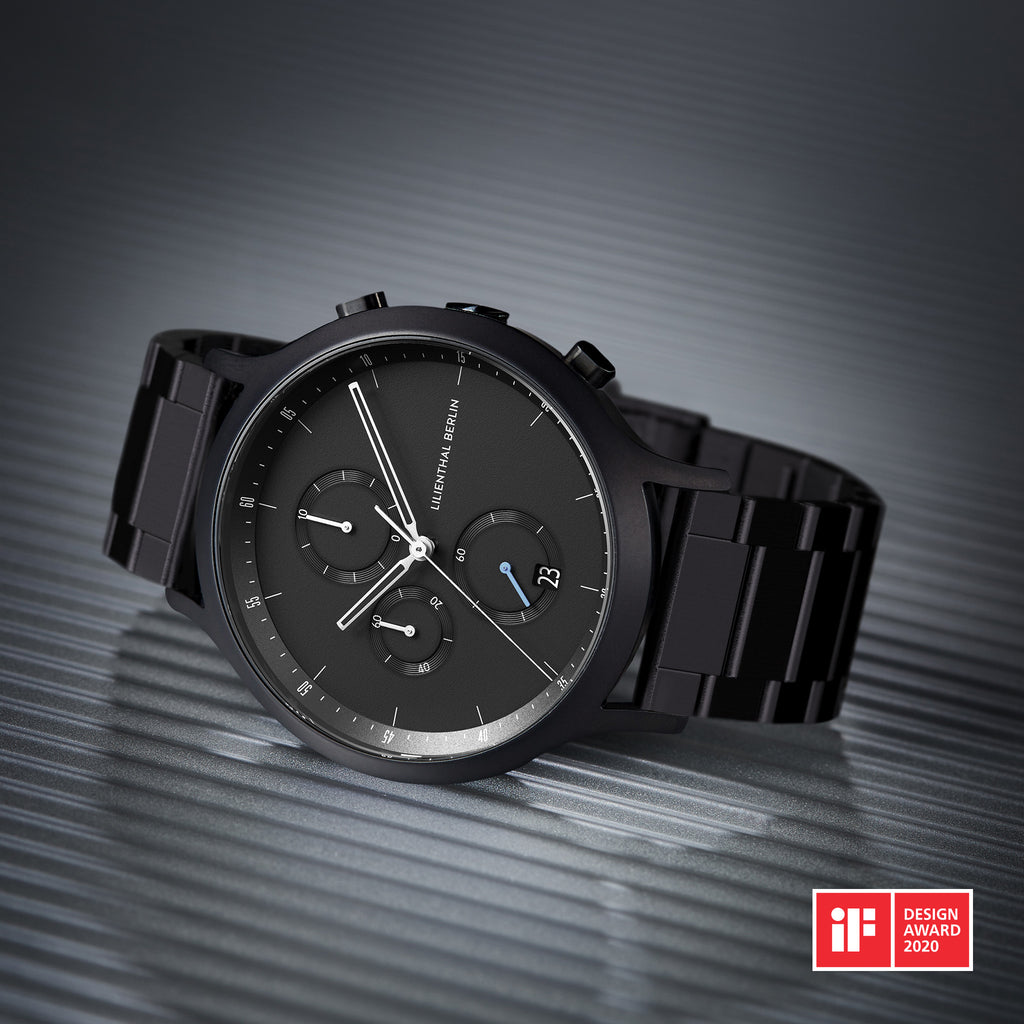Black chronograph watch with metal bracelet on textured surface, featuring date display and minimalist design