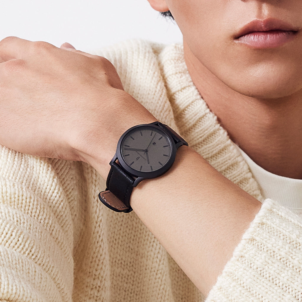 Minimalist black wristwatch with leather strap on wrist, worn over cream knit sweater