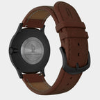 Close-up of the back of a black wristwatch with brown leather strap and engraved caseback.