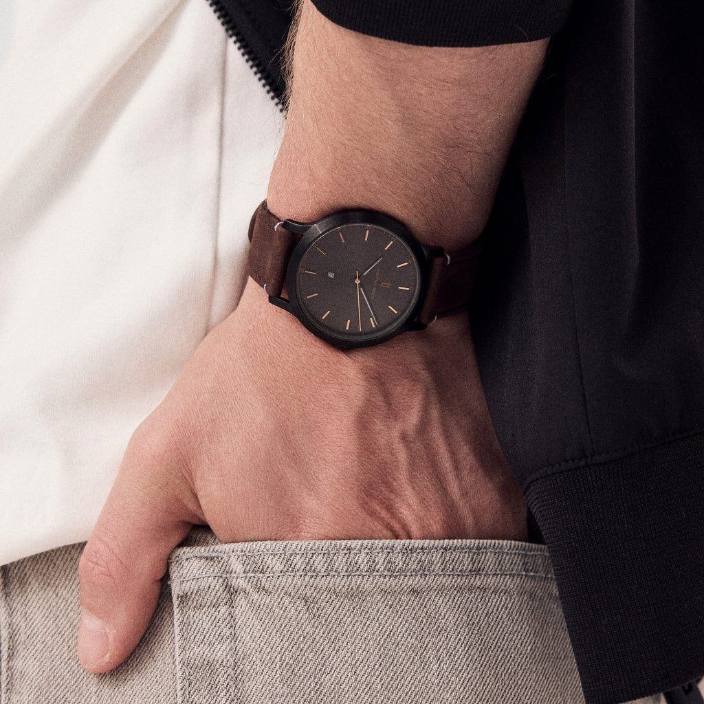 Minimalist black wristwatch with a brown leather strap on a person’s wrist in casual attire