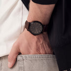 Minimalist black wristwatch with brown leather strap on wrist, casual outfit visible