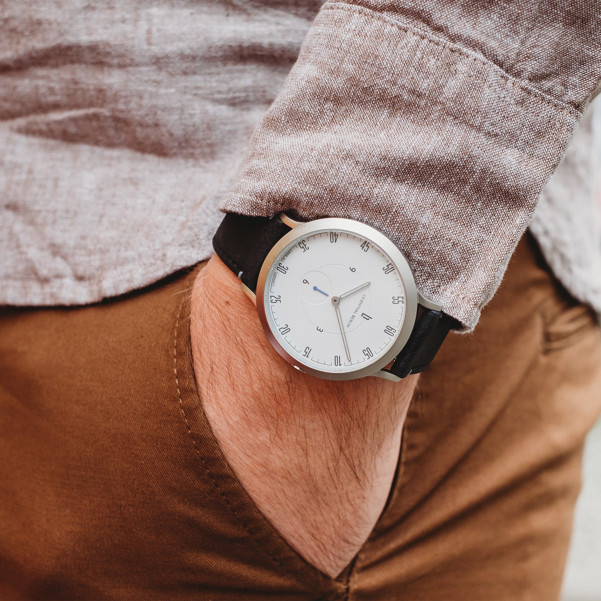 Minimalist silver wristwatch with white dial and black band on wrist, hand in pocket