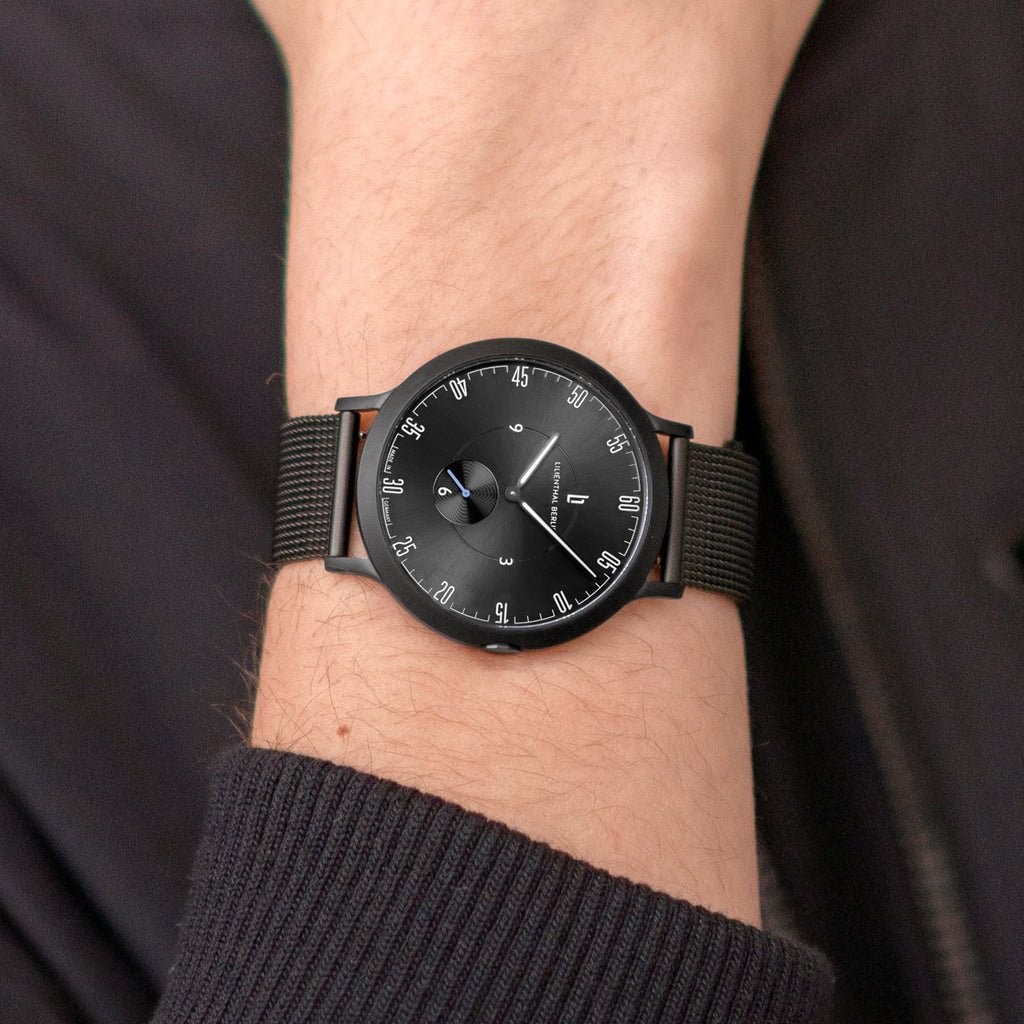 Minimalist black wristwatch with mesh strap on person’s wrist, modern analog dial design